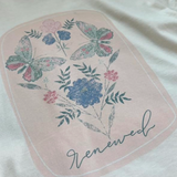 Close-up of “Renewed” butterfly and floral design on soft cream t-shirt, featuring pastel blues, pinks, and greens with delicate script text.